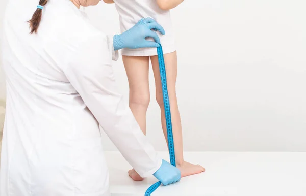 Um médico em luvas azuis com uma fita métrica mede o comprimento das pernas de uma menina caucasiana de 4 anos. Conceito de desenvolvimento e crescimento infantil. Espaço de cópia para texto — Fotografia de Stock