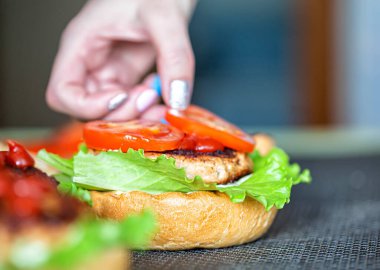 Bir kadının eli lezzetli bir ev yapımı burgerin üzerine bir dilim domates bırakıyor. El yapımı hamburgerler, yakın plan. Hamburger.