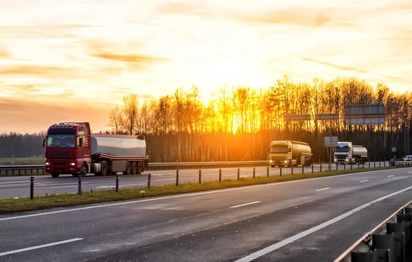 Un convoy de tres camiones transporta mercancías peligrosas en semirremolques cisterna en una autopista con el telón de fondo de la puesta del sol en la noche. —  Fotos de Stock