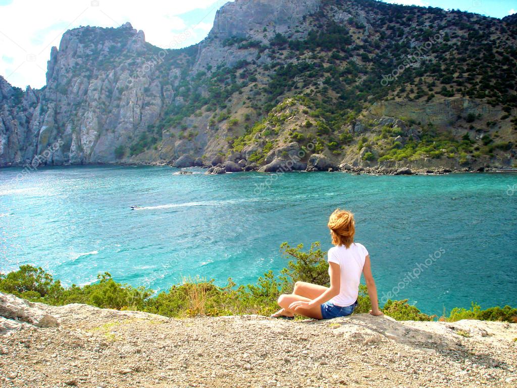 Beautiful blond girl sits on the rock and looks at the sea