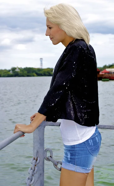 Beautiful blond caucasian model girl in black jacket and jeans on the pier — Stock Photo, Image