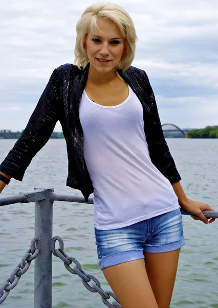 Prachtige blond Kaukasische model meisje in zwarte jas en jeans op de pier — Stockfoto