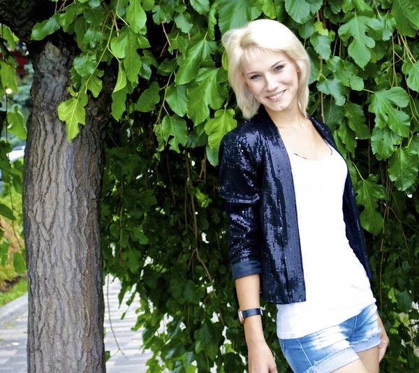 Beautiful blond caucasian model girl poses near the green tree as a backgo — Stock Photo, Image