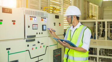 Miğferli bir teknisyen, elektrik kontrol dolabını kontrol ederken ve not alırken elinde bir pano tutuyor..