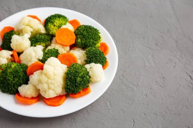 Gri bir yüzey üzerinde, havuç, brokoli ve karnabahar karışımı organik buharda pişirilmiş sebzeler. Metin için boşluk.