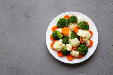 Gri arkaplanda bir plaka üzerinde karışık organik buharda pişirilmiş sebzeler (Havuç, Brokoli ve Karnabahar). Dümdüz, tepeden, yukarıdan. Boşluğu kopyala.