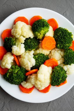 Gri arkaplanda bir plaka üzerinde karışık organik buharda pişirilmiş sebzeler (Havuç, Brokoli ve Karnabahar). Dümdüz, tepeden, yukarıdan.