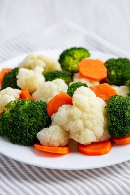 Beyaz ahşap bir zemin üzerinde, havuç, brokoli ve karnabahar karışımı organik buharda pişirilmiş sebzeler..