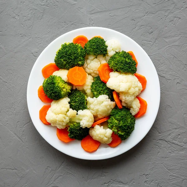 Gri arkaplanda bir plaka üzerinde karışık organik buharda pişirilmiş sebzeler (Havuç, Brokoli ve Karnabahar). Dümdüz, tepeden, yukarıdan. 