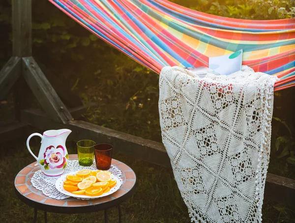 Hammock in garden. Orange, glasses and decanter on small table. — Stock Photo, Image