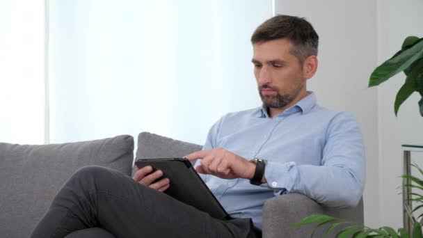 ビジネスマンライトブルーのシャツのソファの上に座っているタブレットテーピング画面を使用して — ストック動画