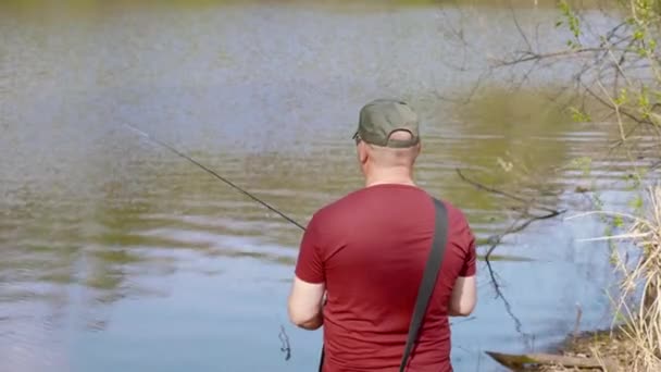 Vista posterior pescador en camiseta roja y la pesca de la tapa mientras que el río de la costa de pie — Vídeos de Stock