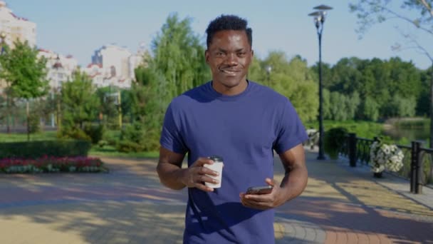 Sonriente hombre afroamericano camina calle utiliza teléfono móvil sosteniendo taza con café — Vídeos de Stock