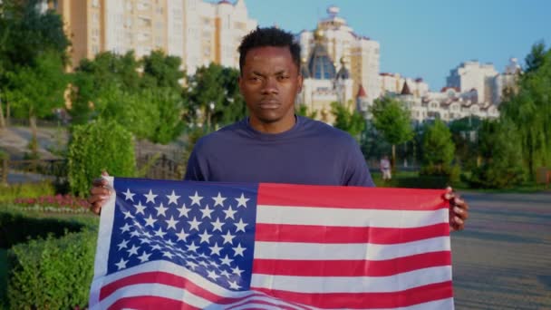 Портрет афро-американца, держащего американский флаг и смотрящего в камеру летом — стоковое видео