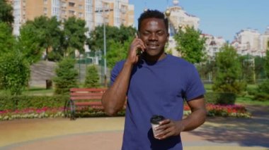 Portre gülümseyen Afro-Amerikalı adam cep telefonuyla konuşuyor. Yazın kahve ya da çay için kağıt bardak tutuyor. Mutlu Afrikalı yetişkin erkek etnik zenci en iyi arkadaşıyla parkta akıllı telefon iletişimi kuruyor