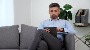 Tablet kullanarak koltukta oturan açık mavi gömlekli bir iş adamı. Evde çalışan serbest çalışan borsacı kanepede oturuyor. Sakallı yetişkin adam tablet ekran görüntüsü kaydediyor