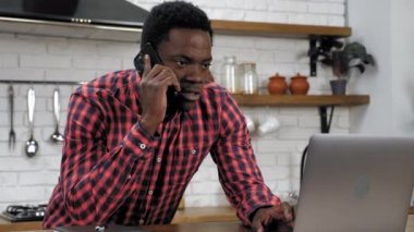 Afrikalı Amerikalı adam dizüstü bilgisayar kullanıyor cep telefonuyla konuşuyor mutfakta duruyor.