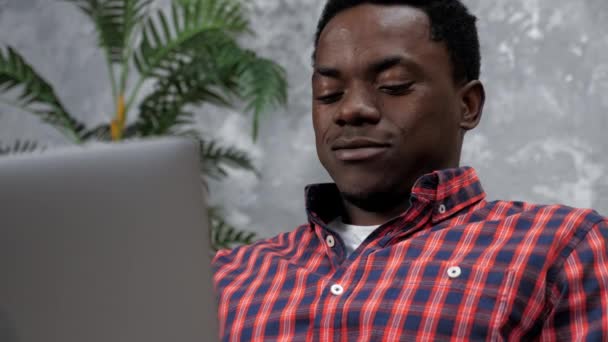 Улыбающийся взрослый афроамериканец, сидящий на диване, работает на ноутбуке, печатая текст — стоковое видео