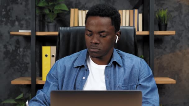 Афроамериканец работает на ноутбук в офисе, пишет информацию в блокноте — стоковое видео