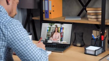 Ofisteki adam internetten video çalışıyor. Bilgisayarı arıyor, öğretmeni dinliyor, defterine yazıyor.