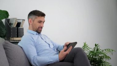 Açık mavi gömlekli gülümseyen işadamı tablet kullanarak koltukta oturuyor.