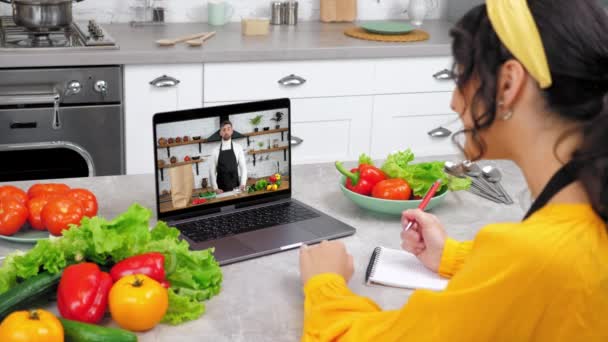 Женщина в кухонном кабинете онлайн видео звонок ноутбук приветствует шеф-повара пишет в блокноте — стоковое видео