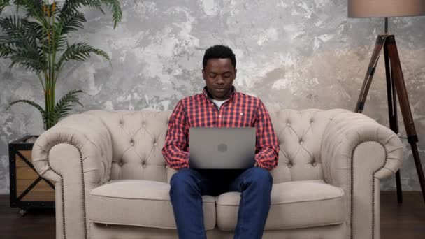 Sonriente hombre afroamericano sentado en el trabajo de sofá para el ordenador portátil escribiendo en el teclado — Vídeo de stock