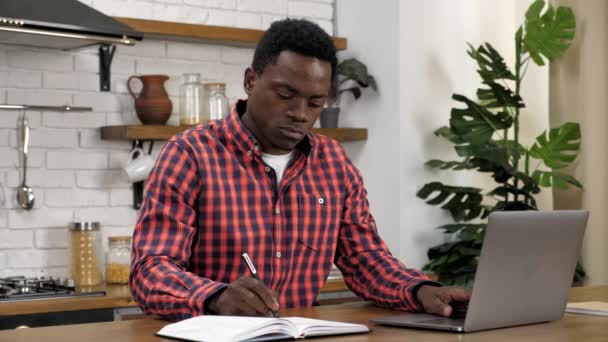 L'uomo afroamericano freelance lavora per computer portatile in cucina, scrive in notebook — Video Stock