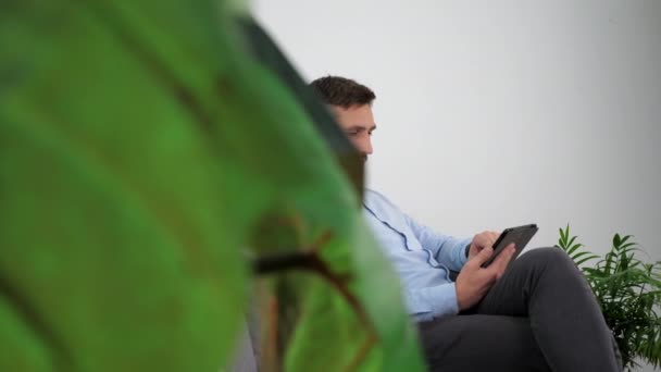 Улыбающийся бизнесмен в светло-голубой рубашке сидит на диване с помощью планшета — стоковое видео
