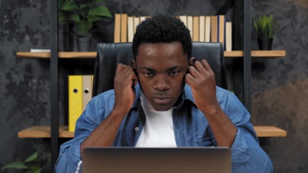 Афроамериканец вставляет беспроводные наушники в уши, работает на ноутбук — стоковое видео