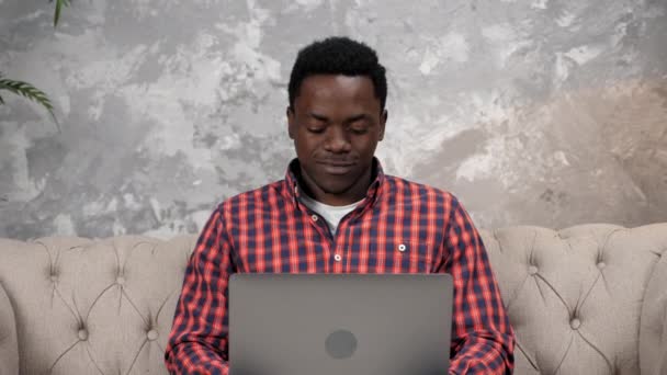 Sonriente hombre afroamericano sentado en el trabajo de sofá para el ordenador portátil escribiendo en el teclado — Vídeo de stock