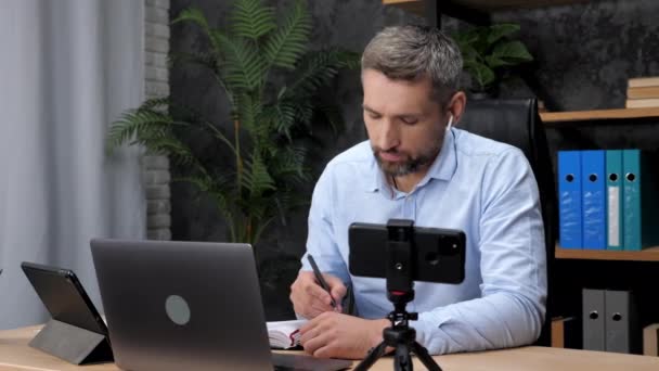 Homme commerçant communique ordinateur portable en ligne avec l'investisseur, prend des notes dans le carnet — Video