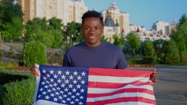 Portrait homme afro-américain tenant un drapeau américain et regarde la caméra en été — Video