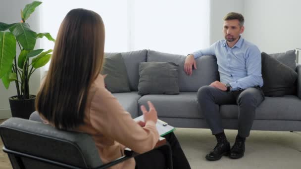 Дорослий бородатий чоловік з проблемами психічного здоров'я сидить на дивані слухати терапевта — стокове відео