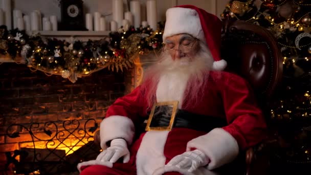 Santa Claus falls asleep sitting on chair on background fireplace Christmas tree — Stock Video