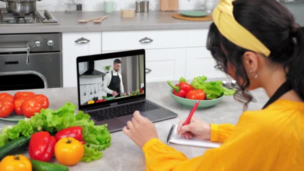 Mulher na cozinha estudar aula de culinária remota ouvir chef, escreve em notebook — Vídeo de Stock