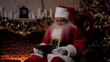 Modern gülümseyen Noel Baba, yeni yıl kış tatili tabletinde mobil oyun oynar.