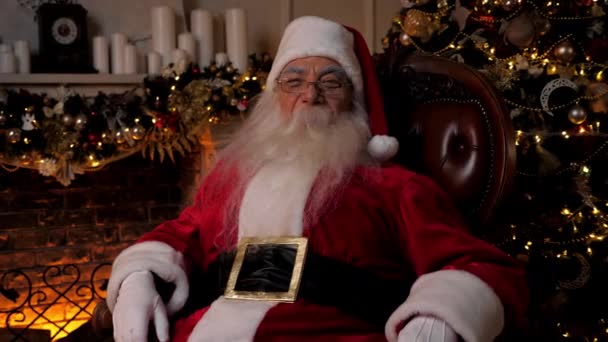 Santa Claus sonriente se ve la cámara sentado en la silla en casa. Vacaciones de Navidad — Vídeos de Stock