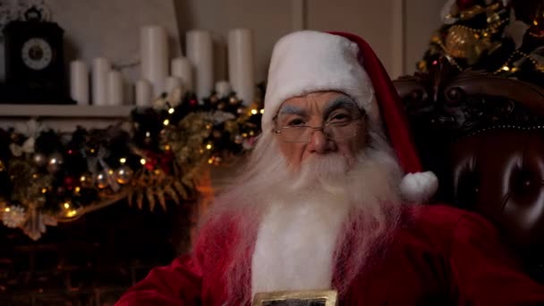 Κοντινό πλάνο Άγιος Βασίλης κοιτάζει κάμερα κάθεται στην καρέκλα στο σπίτι, Χριστούγεννα διακοπές — Αρχείο Βίντεο