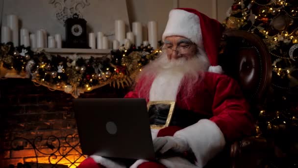 El viejo Papá Noel sonriente moderno utiliza los rellenos del ordenador portátil en tarjetas de vacaciones para los niños — Vídeos de Stock