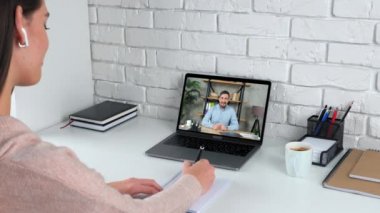 Evde ders çalışan bir kadın internetten interneti izler, öğretmenini dinler, defterine yazar.