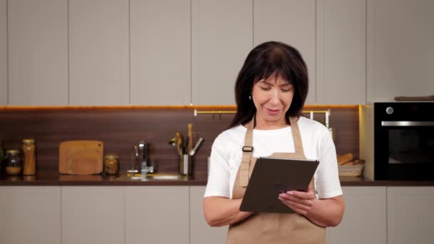 Smiling aged woman uses tablet notes list ingredients for dish in home kitchen — Vídeo de Stock