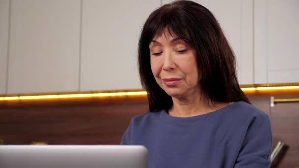 Mujer anciana sonriente utiliza la escritura de computadoras portátiles en el teclado en la cocina casera, trabajo a distancia — Vídeos de Stock