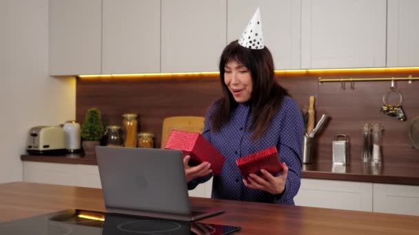 Aged woman in party hat opens red gift box and is surprised in home kitchen — Stock Video