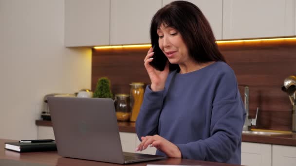 Mulher de negócios sorrindo falando no telefone celular funciona para laptop na cozinha doméstica — Vídeo de Stock