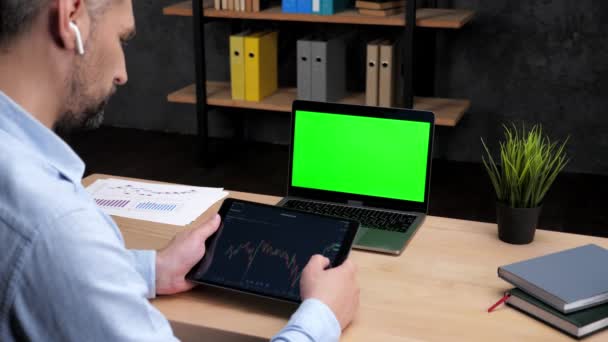 Man broker scrollen tablet met aandelenbeurs grafiek, laptop met groen scherm — Stockvideo