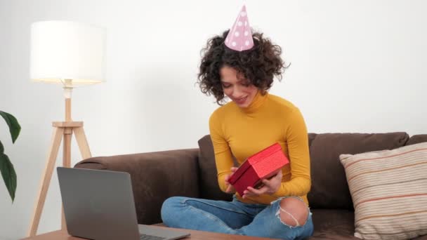 Happy woman in party hat opens gift box and surprised, sitting in couch at home — ストック動画