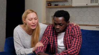 Caucasian woman calms African American upset man sitting on couch at home