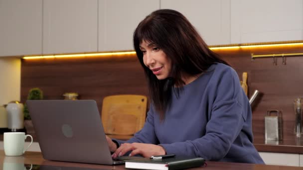 Smiling aged woman housewife uses laptop typing text on keyboard in home kitchen — Stock Video