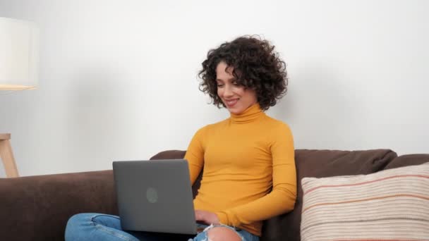 Smiling curly woman student uses laptop typing on keyboard sitting on couch — ストック動画